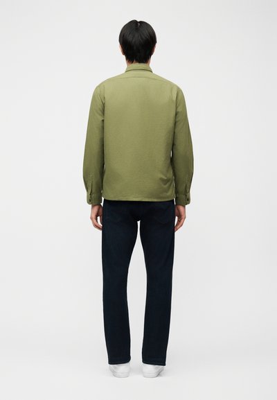 Άτομο που στέκεται με την πλάτη γυρισμένη, φορώντας πουκάμισο χακί με κουμπιά, σκούρο τζιν και λευκά αθλητικά παπούτσια σε λευκό φόντο.