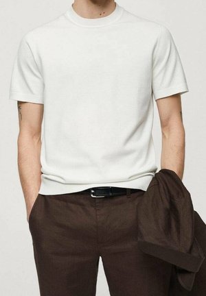 T-shirt blanc à manches courtes et col rond en tissu texturé, associé à un pantalon marron et une veste assortie tenue sur le bras.