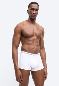 Μυώδης άνδρας όρθιος, φορώντας λευκά μποξεράκια με το λογότυπο GANT, μπροστά σε απλό ανοιχτό γκρι φόντο.