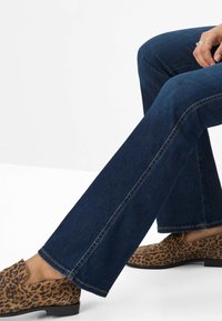 Jeans a zampa d'elefante blu scuro, aderenti, con cuciture visibili, abbinati a mocassini con stampa leopardata su uno sfondo uniforme.
