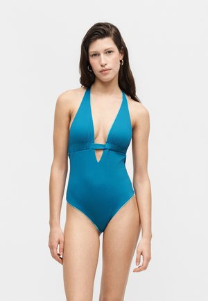 Femme portant un maillot de bain une-pièce bleu sarcelle à col halter, avec une découpe en V profonde et une petite découpe sous la poitrine, debout devant un fond uni.