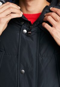 Černá prošívaná bunda s kapucí, zapínání na zip, druky a červený svetr pod ní. Texturovaná látka s viditelným prošíváním.