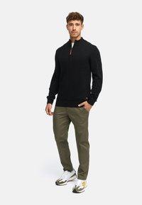 Czarny sweter z zamkiem w serek o prążkowanej teksturze, połączony z khaki spodniami i białymi trampkami z kamuflażowymi akcentami, tworząc zsynchronizowany outfit.