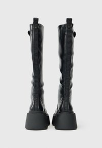 Crne čizme do koljena od teksturirane kože. Robustan kvadratni potplat sa utisnutim logotipom sa strane. Glatka površina s trakama za povlačenje.
