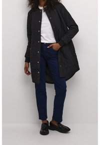 Schwarze gesteppte lange Jacke mit rundem Kragen, Druckknöpfen und Seitentaschen, kombiniert mit dunkelblauen Jeans und schwarzen Loafers.