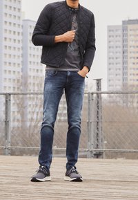 Schwarze gesteppte Bomberjacke über einem grauen Grafik-T-Shirt, kombiniert mit blauen Jeans und schwarzen Sportschuhen mit weißen Sohlen.