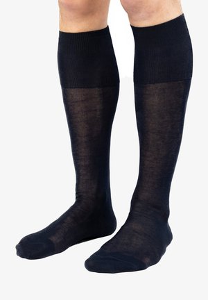 Beine, die kniehohe, halbtransparente schwarze Socken mit einem festen gerippten Bündchen tragen, vor einem schlichten weißen Hintergrund stehend.
