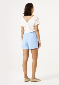 Haut blanc à manches courtes avec un dos en V, associé à un short bleu clair. Le mannequin porte des ballerines tissées beiges. Design simple, textures douces.