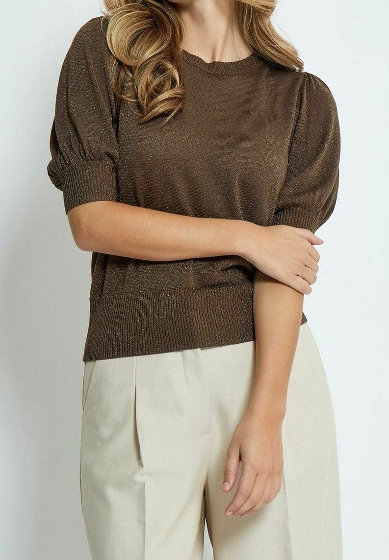 Femme portant un haut marron à manches courtes bouffantes et un pantalon plissé crème taille haute, debout les bras croisés.