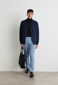Marine blå strikket cardigan, svart turtleneck, mønstrede lysblå bukser, og svart veske. Modellen har på seg svarte sko og står mot en nøytral bakgrunn.