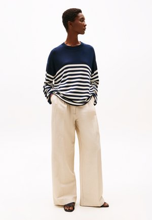 Oseba, oblečena v mornarsko modro in belo črtasto jopico, bež široke hlače in črne sandale, stoji z rokami v žepih, obrnjena proti desni.