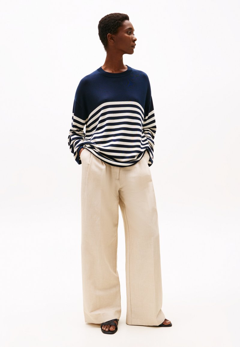 Person iført en navy- og hvidstribet sweater, beige bukser med vide ben og sorte sandaler, stående med hænderne i lommerne, vendt mod højre.