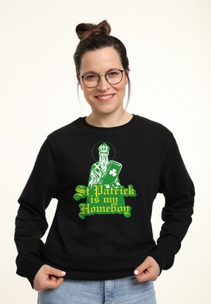 Smilende kvinde med briller iført sort sweatshirt med motiv af Sankt Patrick, der holder en tablet med en kløver, og teksten "St. Patrick er min ven."