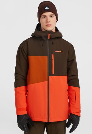 Bruine en oranje waterdichte jas met capuchon, met een kleurblokontwerp, een voorkantsluiting en zijzakken. Zwarte handschoenen gedragen.