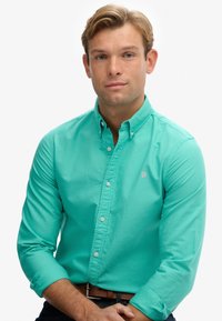 Mintgroene blouse met een kraag, lange mouwen opgerold en een klein embleem op de borst. Draagt een bruine riem en donkere jeans.