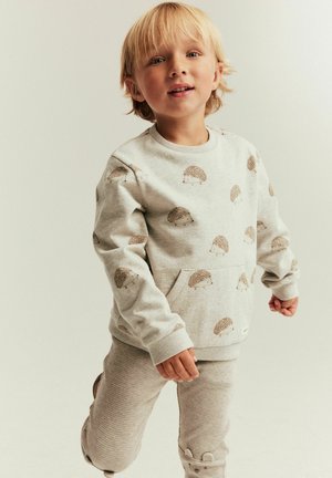 Jong kind met blond haar, gekleed in een beige trui met egelprint en beige textuurbroek, staand tegen een neutrale achtergrond.