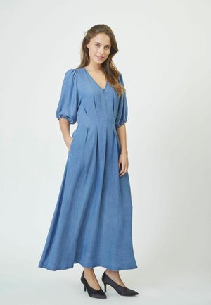 Denim maxi dress with short puff sleeves, V-neckline, pleats, and pockets. Light blue fabric, paired with black pointed high heels.