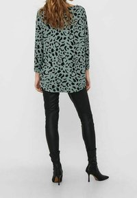 Langärmliges, mintgrünes Oberteil mit schwarzem Leopardenmuster, kombiniert mit figurbetonten schwarzen Lederhosen und spitzen, schwarzen High Heels.