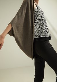 Lekki brązowy kardigan na czarno-białej wzorzystej bluzce, zestawiony z czarnymi dopasowanymi spodniami. Miękka tekstura i luźny krój.