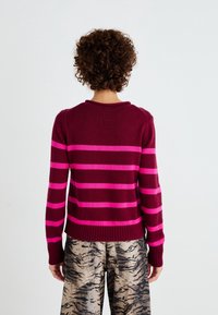 Personne aux cheveux bouclés portant un pull bordeaux avec des rayures horizontales roses et un pantalon beige ample avec des motifs abstraits noirs, vue de dos.