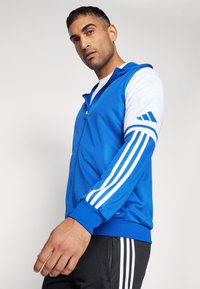 Blå zip-up hettegenser med hvite ermer som har tre vertikale striper. Materialet ser glatt ut; inkluderer detaljer på mansjettene og logo.