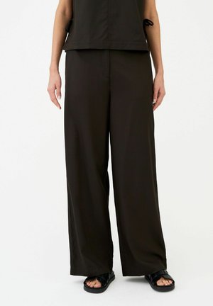 Femme portant un pantalon large noir ample, un haut noir sans manches et des sandales noires ouvertes à l'avant, debout devant un fond blanc uni.