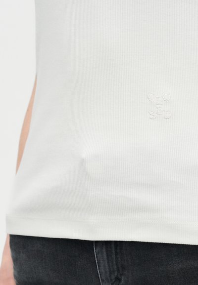 Gros plan sur un t-shirt blanc à côtes avec un petit texte brodé en blanc "CLOSED" sur le devant, porté avec un jean en denim foncé.