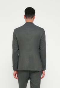Man met kort krullend haar in een slim-fit donkergrijs pak, staand met het gezicht naar een eenvoudige witte achtergrond, met ontspannen armen langs zijn zij.
