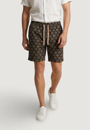 Uomo che indossa pantaloncini neri con motivo e coulisse rosa, una camicia bianca a maniche corte abbottonata e scarpe da ginnastica bianche che cammina in avanti.