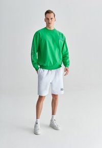 Zielona bluza z okrągłym dekoltem i graficznym napisem na rękawie, w połączeniu z białymi szortami z haftowanym logo. Całość dopełniają białe trampki.