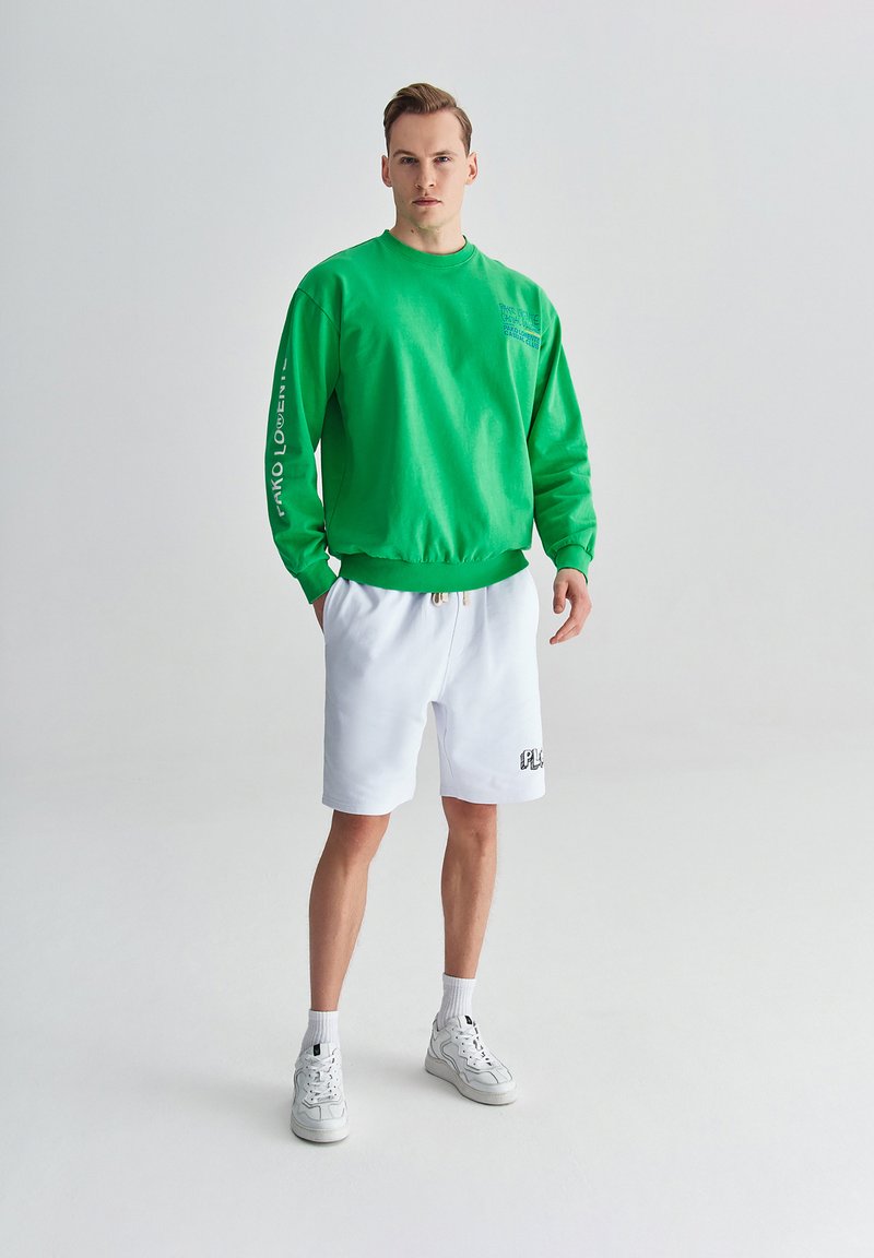 Zielona bluza z okrągłym dekoltem i graficznym napisem na rękawie, w połączeniu z białymi szortami z haftowanym logo. Całość dopełniają białe trampki.