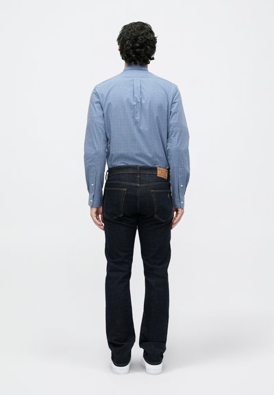 Mėlynos langelinės marškiniai su ilgomis rankovėmis, derinami su tamsaus džinsinio audinio kelnėmis ir baltais sportbačiais, matomi iš nugaros.