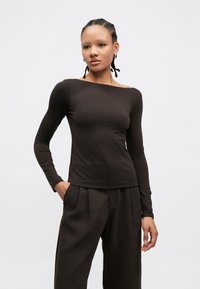 Τοπ με μακριά μανίκια και ανοιχτούς ώμους σε σκούρο καφέ ύφασμα, με εφαρμοστή γραμμή και λεία υφή, συνδυασμένο με ψηλόμεσο σκούρο παντελόνι.