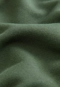 Tejido verde con una superficie lisa y ligeramente texturizada. El material parece suave y ligero, mostrando un patrón de punto consistente.