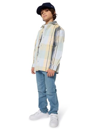 Garçon portant une chemise à carreaux claire et à boutons dans des couleurs pastel, un jean bleu et des baskets blanches ; agrémenté d'une casquette marine.