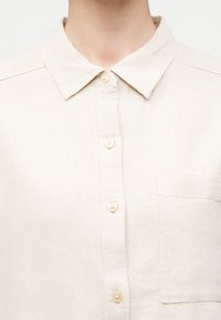 Κοντινό πλάνο ενός ατόμου που φοράει μια μπεζ πουκάμισο με κουμπιά, γιακά και τσέπη στο στήθος στη δεξιά πλευρά.