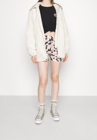 Jasno-beżowa kurtka zapinana na zamek, czarny top z węzłem, szorty w kwiatowy wzór w różu i czerni oraz oliwkowe trampki za kostkę.