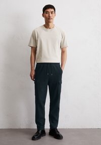 T-shirt en coton beige associé à un pantalon cargo vert foncé avec deux poches latérales, porté avec des bottes noires à lacets.