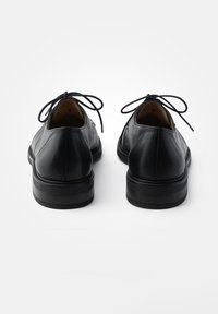 Scarpe in pelle nera con punte rotonde, suole piatte e lacci legati sul retro, caratterizzate da una texture liscia e un design minimalista.