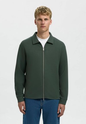 Zeleno zip-up jakna s izraženim ovratnikom, dugim rukavima i glatkom teksturom, nosi se preko bijele majice i uparen je s plavim trapericama.