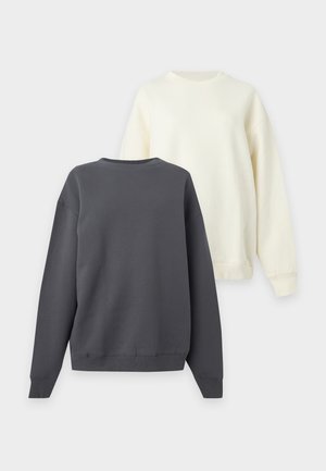 Deux modèles portant des sweat-shirts blancs et des pantalons de survêtement gris avec des bandes blanches sur les côtés, posant devant un fond blanc uni.