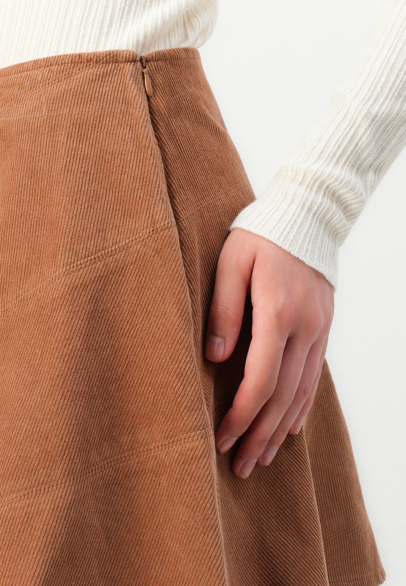 Mano appoggiata su una gonna in corduroy marrone con cerniera visibile, indossando una maglietta a maniche lunghe bianca e a coste, su uno sfondo neutro.