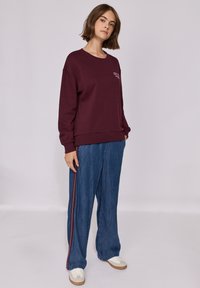 Sweatshirt bordeaux avec un col rond et des poignets côtelés, associé à un pantalon en denim ample avec une bande latérale rouge et blanche, porté avec des baskets blanches.