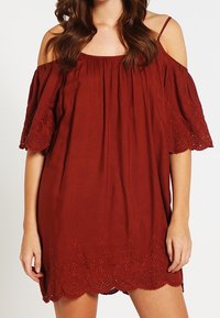 Kastanjefärgad off-shoulder klänning med korta fransade ärmar. Har intrikata spetsdetaljer vid nederkanten och en lös, flytande design.