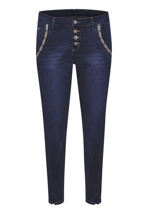 Jean slim bleu foncé avec boutons décoratifs, bordure de poche à motifs, fentes avant aux chevilles, porté avec des baskets blanches et un chemisier blanc à manches longues.