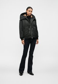 Musta tikattu takki suurella hupulla, ribatulla kauluksessa ja turkisliningillä. Sisältää kaksi etupuuttavaa vetoketjupussia ja vetoketjusulun, yhdistettynä tummiin farkkuihin.