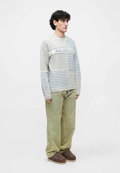 Szürke és kék mintás pulóver kábelkötéssel és csíkokkal, világoszöld bő szárú nadrággal és barna bebújós cipővel párosítva.