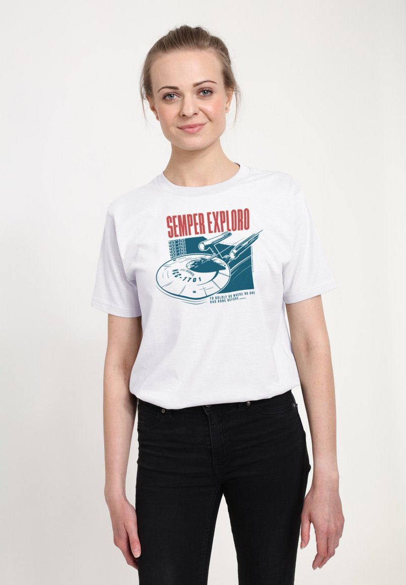 Kvinde iført hvid T-shirt med rumskibs grafik og teksten "SEMPER EXPLORO", kombineret med sorte bukser, stående foran en ensfarvet baggrund.