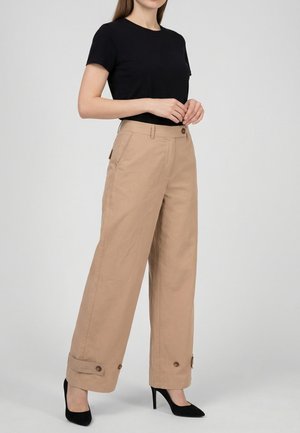 Femme portant un haut noir à manches courtes, un pantalon large beige avec des poignets à boutons, et des chaussures à talons hauts noires, sur un fond uni.