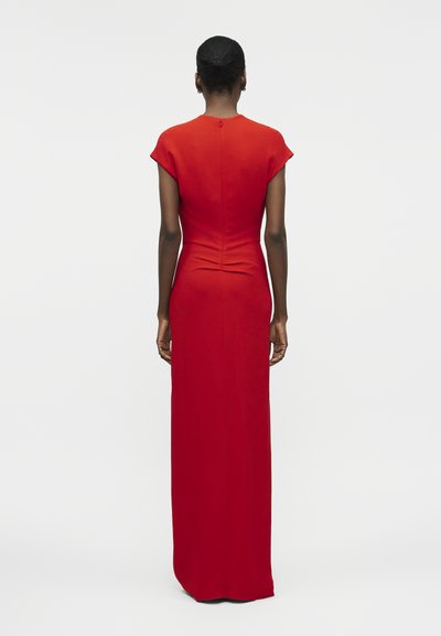 Mujer de cabello corto viste un vestido rojo ajustado hasta el suelo con mangas tipo capa, visto desde atrás sobre un fondo blanco liso.